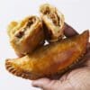 Hand holding two golden-brown fried empanadas, one cut open showing savory filling.