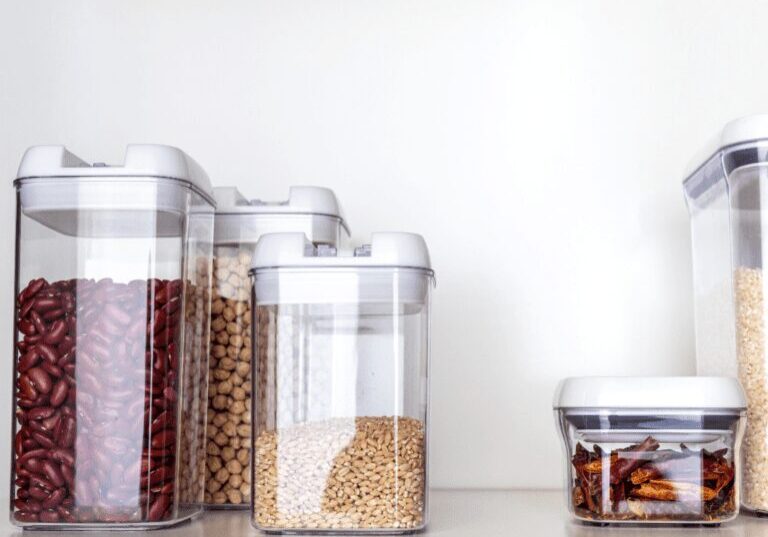 Clear containers hold various dry foods on a white surface against a white wall.