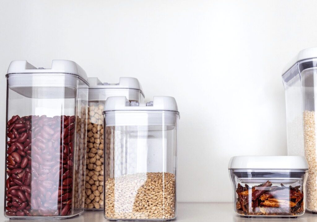 Clear containers hold various dry foods on a white surface against a white wall.