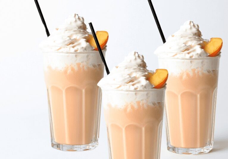 Three peach milkshakes topped with whipped cream and garnished with peach slices.
