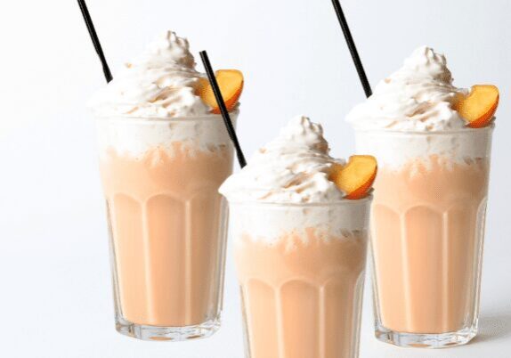 Three peach milkshakes topped with whipped cream and garnished with peach slices.