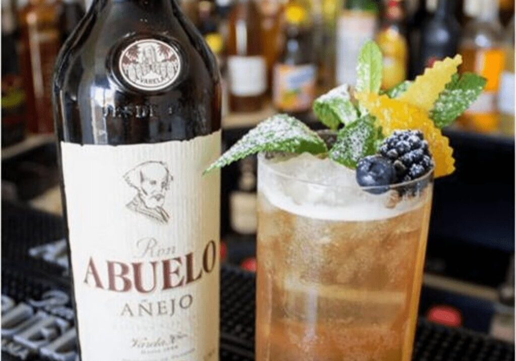 Cocktail garnished with mint, berries, and citrus, beside Abuelo Añejo rum bottle.